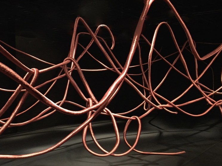 Copper-tinted sculptural cables forming abstract loops against a black background by Alice Anderson.