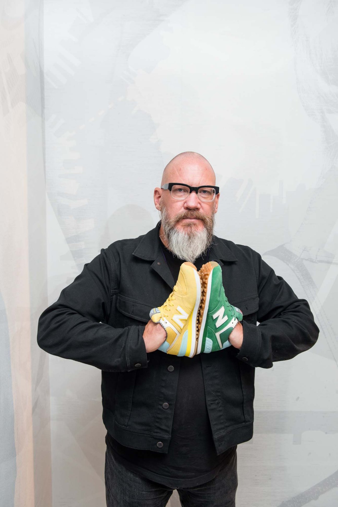 Bob Neville, New Balance's global retail creative director, holds yellow and green New Balance sneakers while wearing glasses and a black jacket.