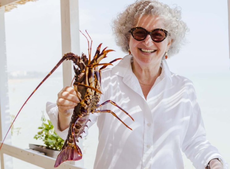 Anissa Helou holds a fresh red lobster while smiling at a seaside restaurant in Sicily.