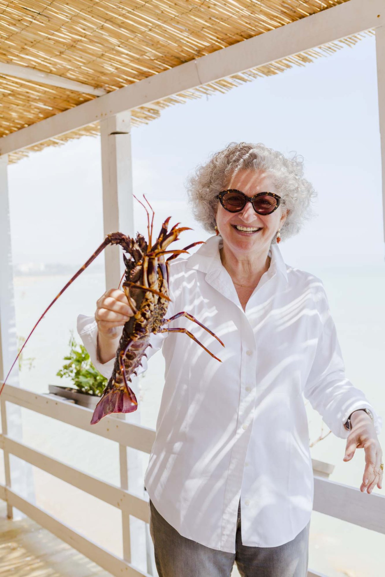 Anissa Helou holds a fresh red lobster while smiling at a seaside restaurant in Sicily.
