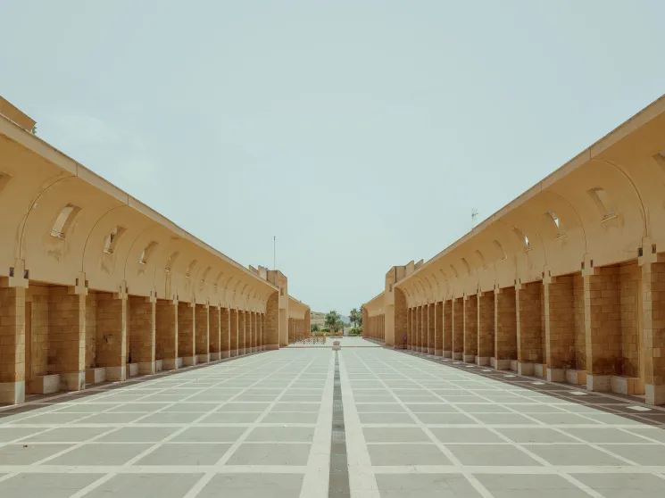 Symmetrical sand-colored stone colonnade with arched openings flanking a geometric paved courtyard under pale sky