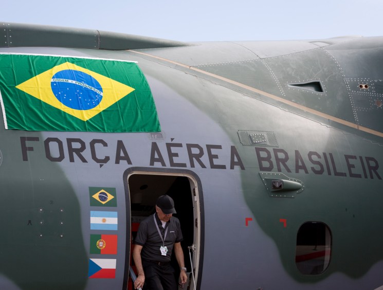 Brazil's Força Aérea Brasileira KC-390 military transport aircraft with Brazilian flag and crew member visible at open door.