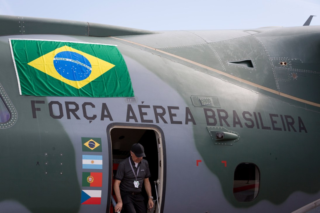Brazil's Força Aérea Brasileira KC-390 military transport aircraft with Brazilian flag and crew member visible at open door.
