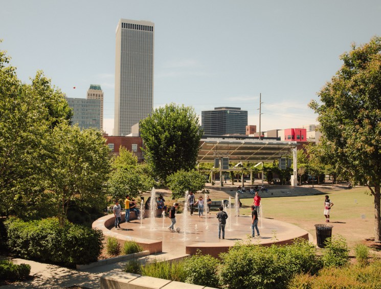 Guthrie Green plaza with fountain, trees, and Tulsa skyline with tall buildings in background.