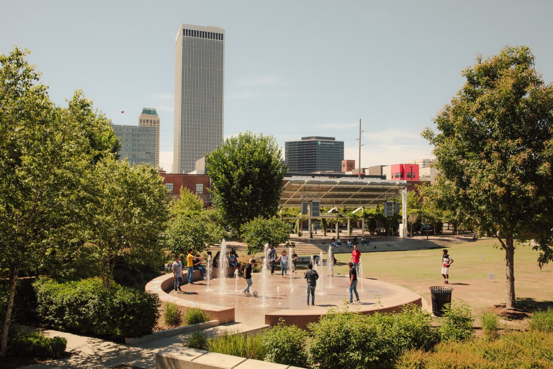 Guthrie Green plaza with fountain, trees, and Tulsa skyline with tall buildings in background.