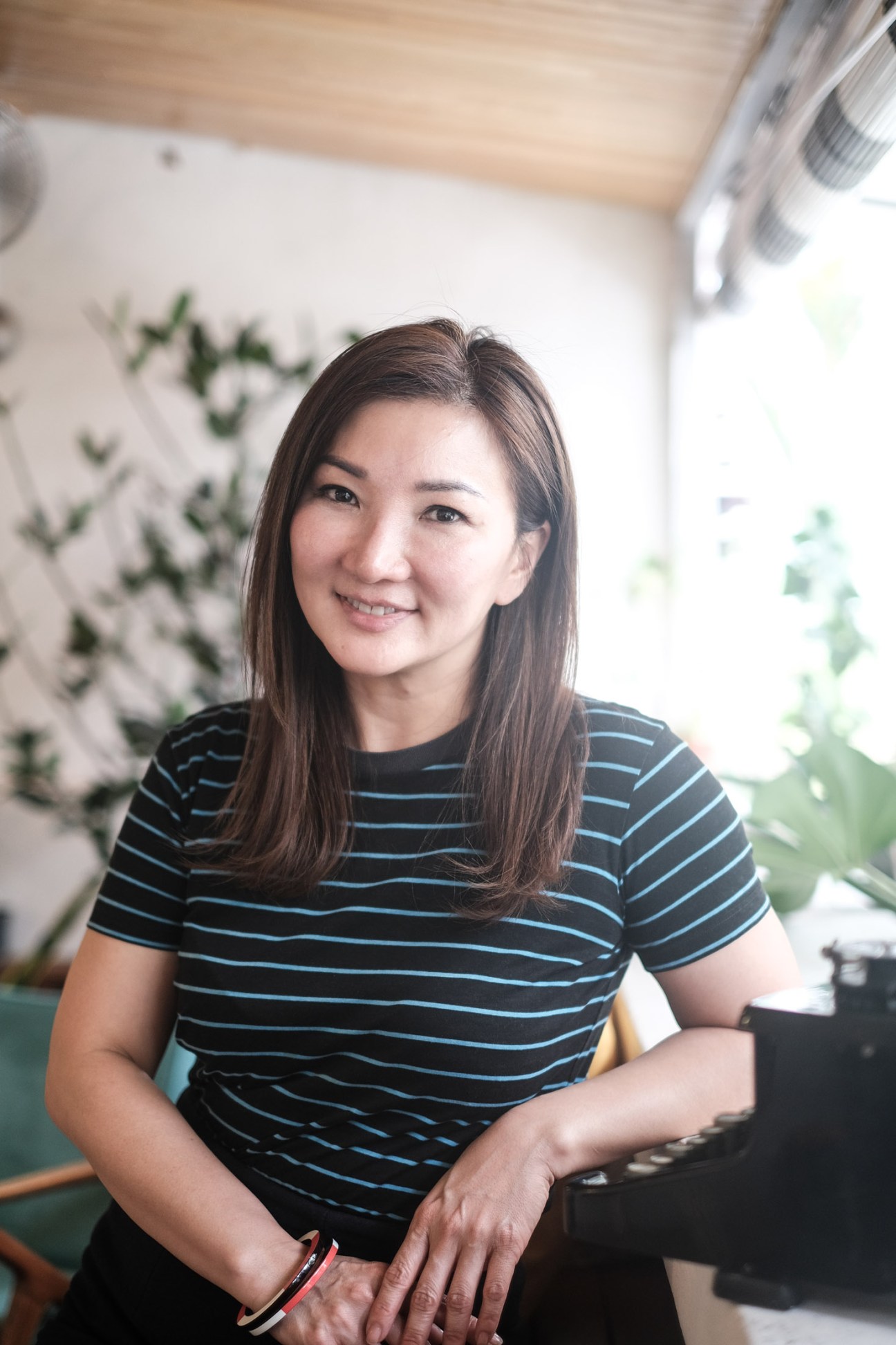 Cynthia Chua, Singaporean beauty and hospitality entrepreneur, smiling in a striped shirt in a bright, plant-filled space.