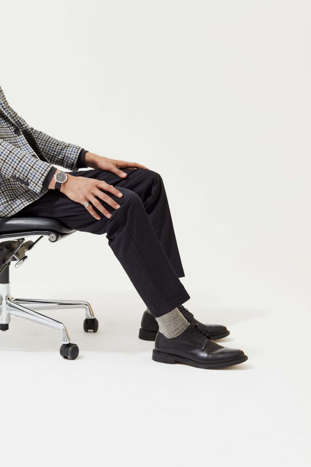 Man seated in office chair wearing checked jacket, black trousers, Church's shoes and Chopard watch