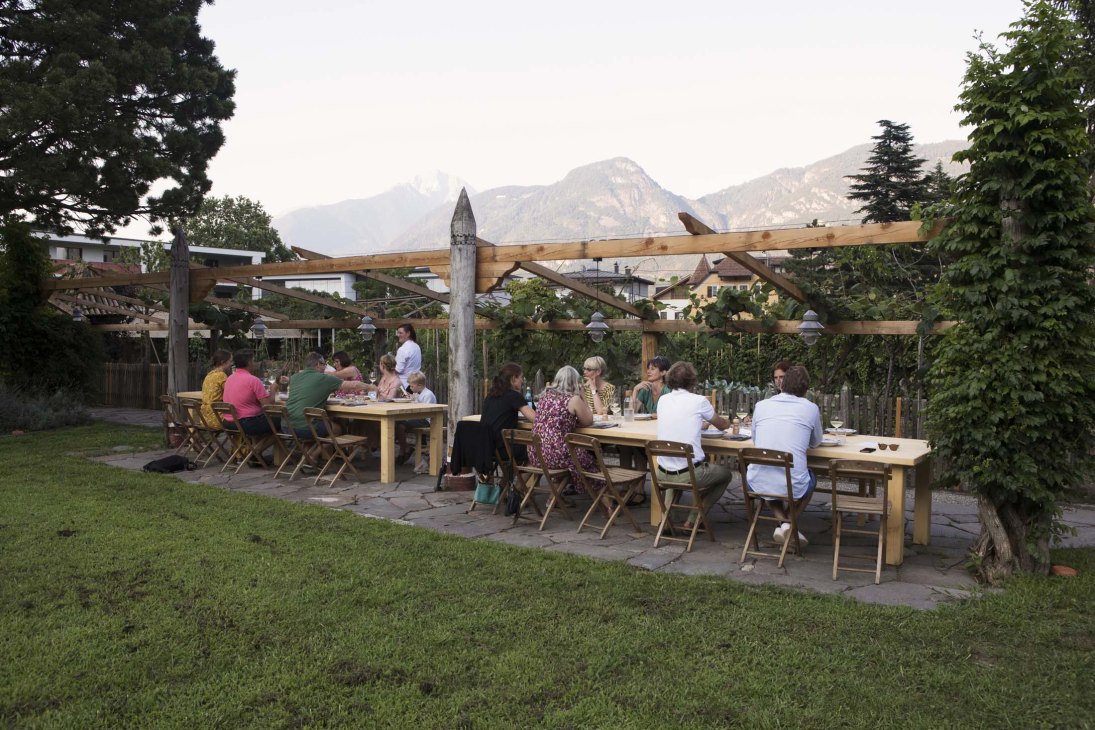 Guests dining at communal tables under a wooden pergola in Villa Arnica's garden with mountain views.