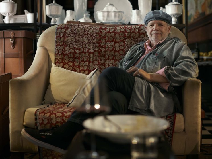 Argentine chef Francis Mallmann seated in a cream armchair in his restaurant, wearing a grey jacket and cap.