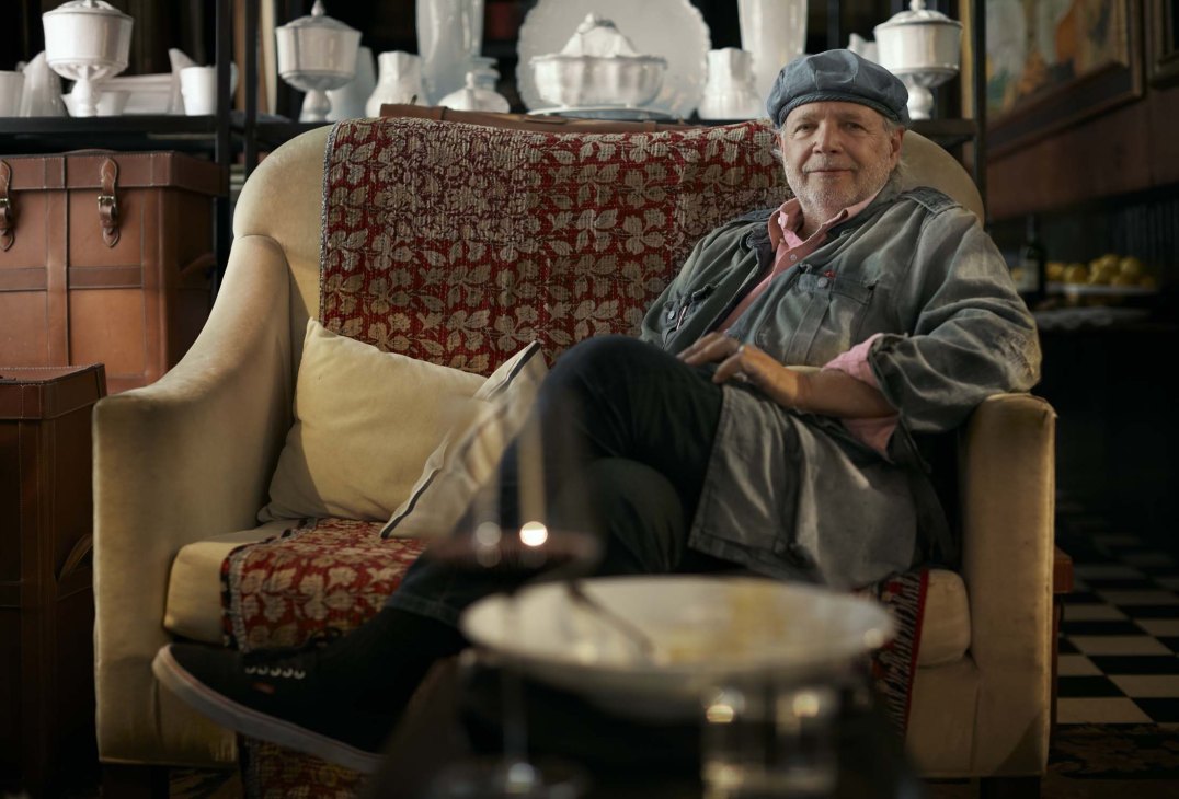 Argentine chef Francis Mallmann seated in a cream armchair in his restaurant, wearing a grey jacket and cap.