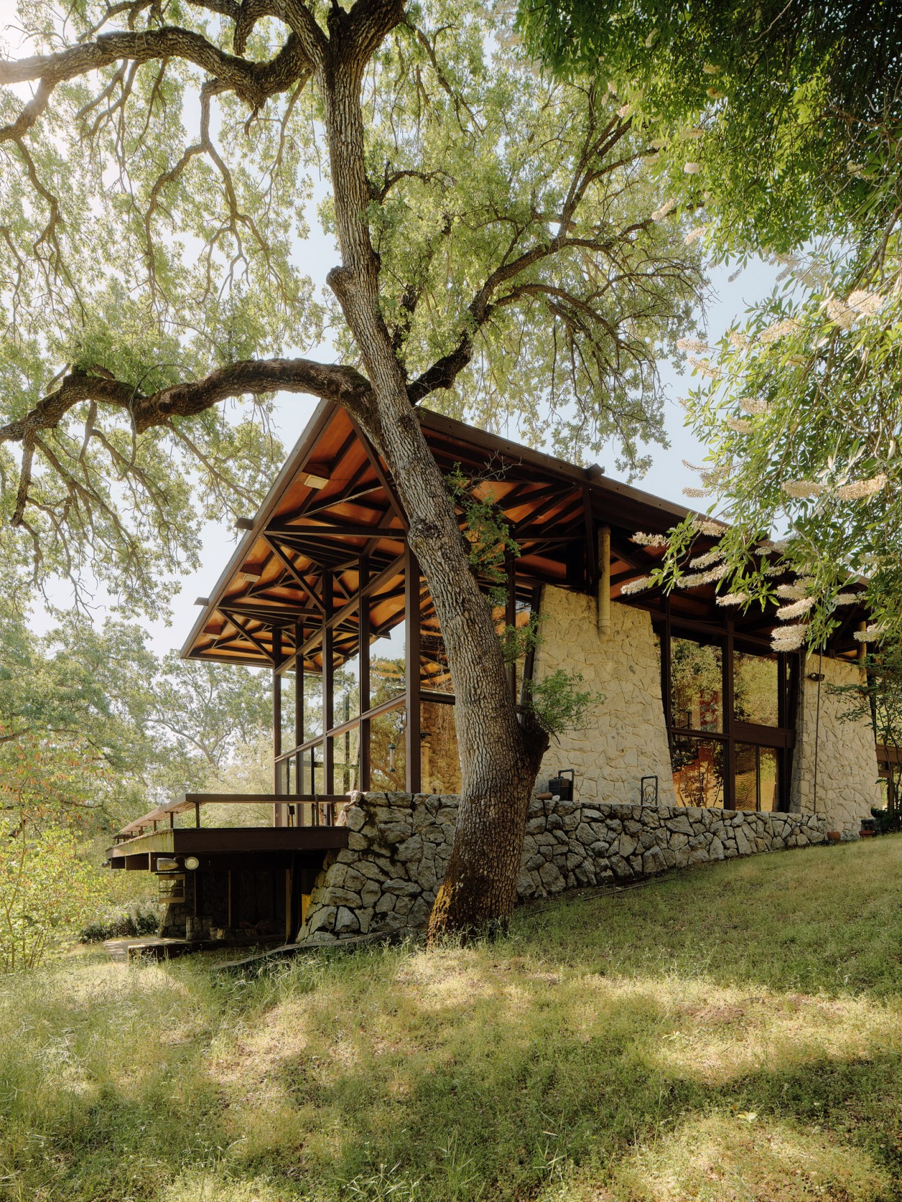 Modernist glass house with wooden beam canopy and stone foundation nestled among oak trees in natural landscape.