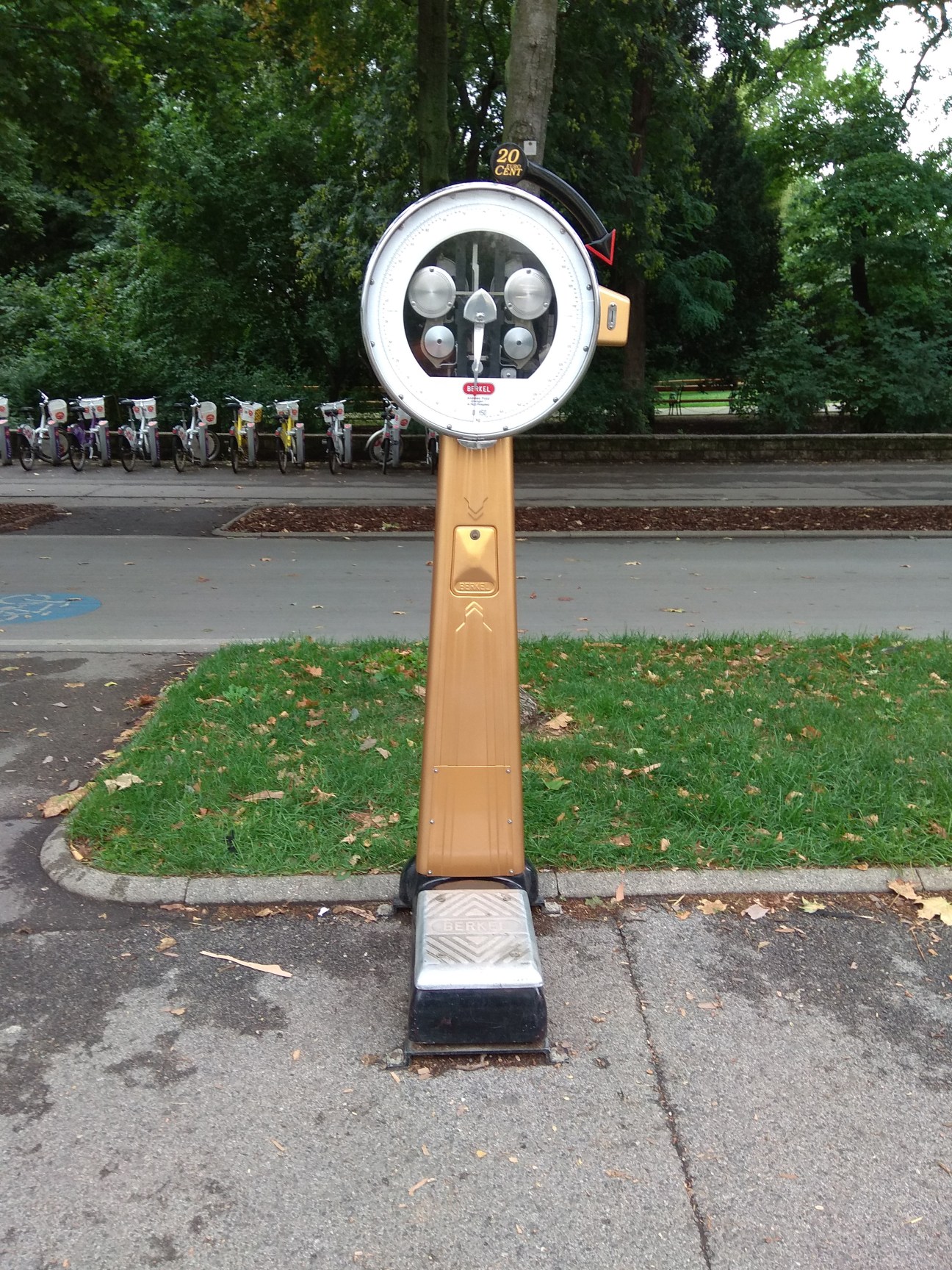 A vintage weighing scale on a sidewalk, with bicycle parked across the road.