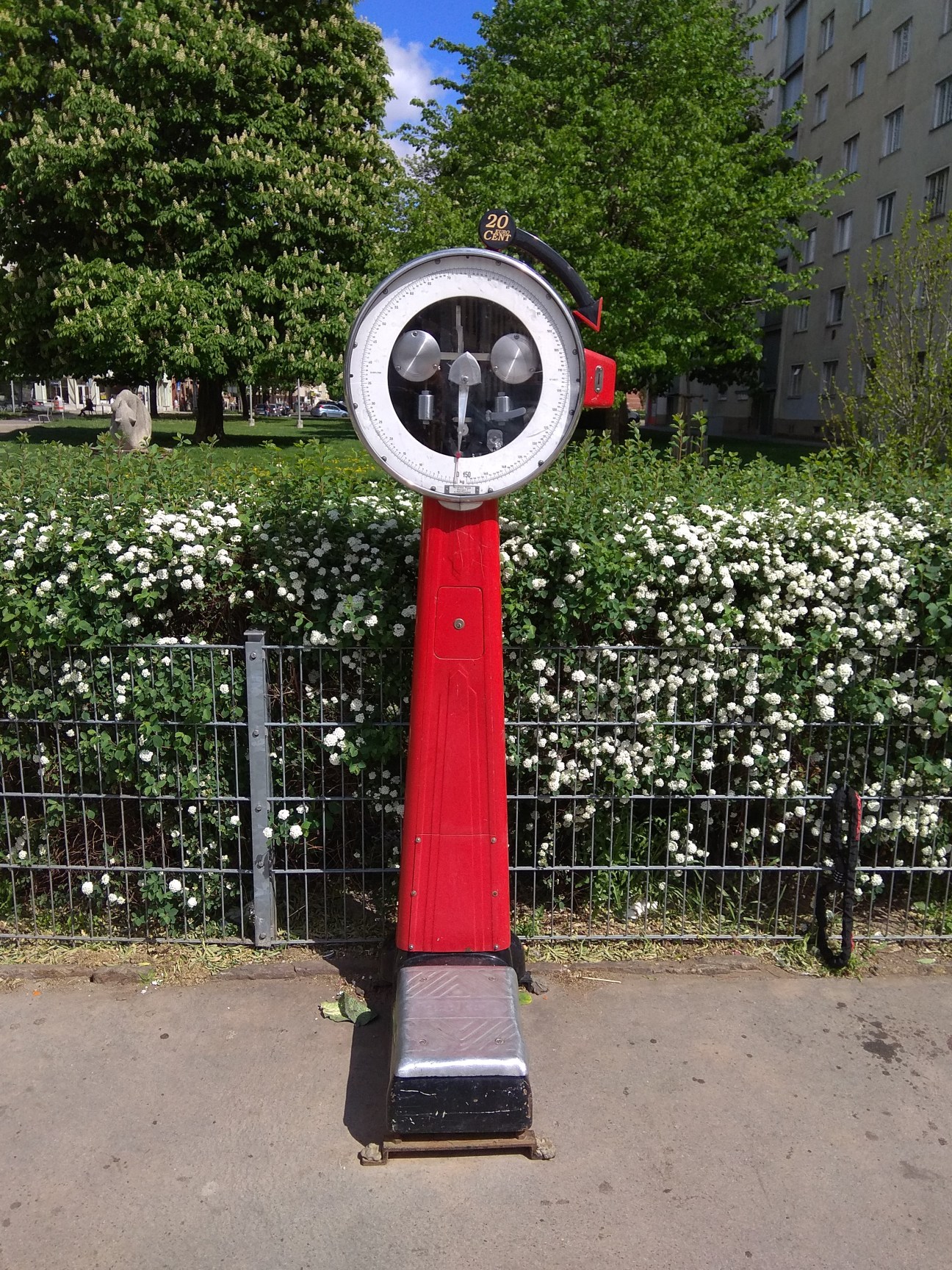 A vintage weighing scale in red and silver, set against a backdrop of green trees and white flowers.