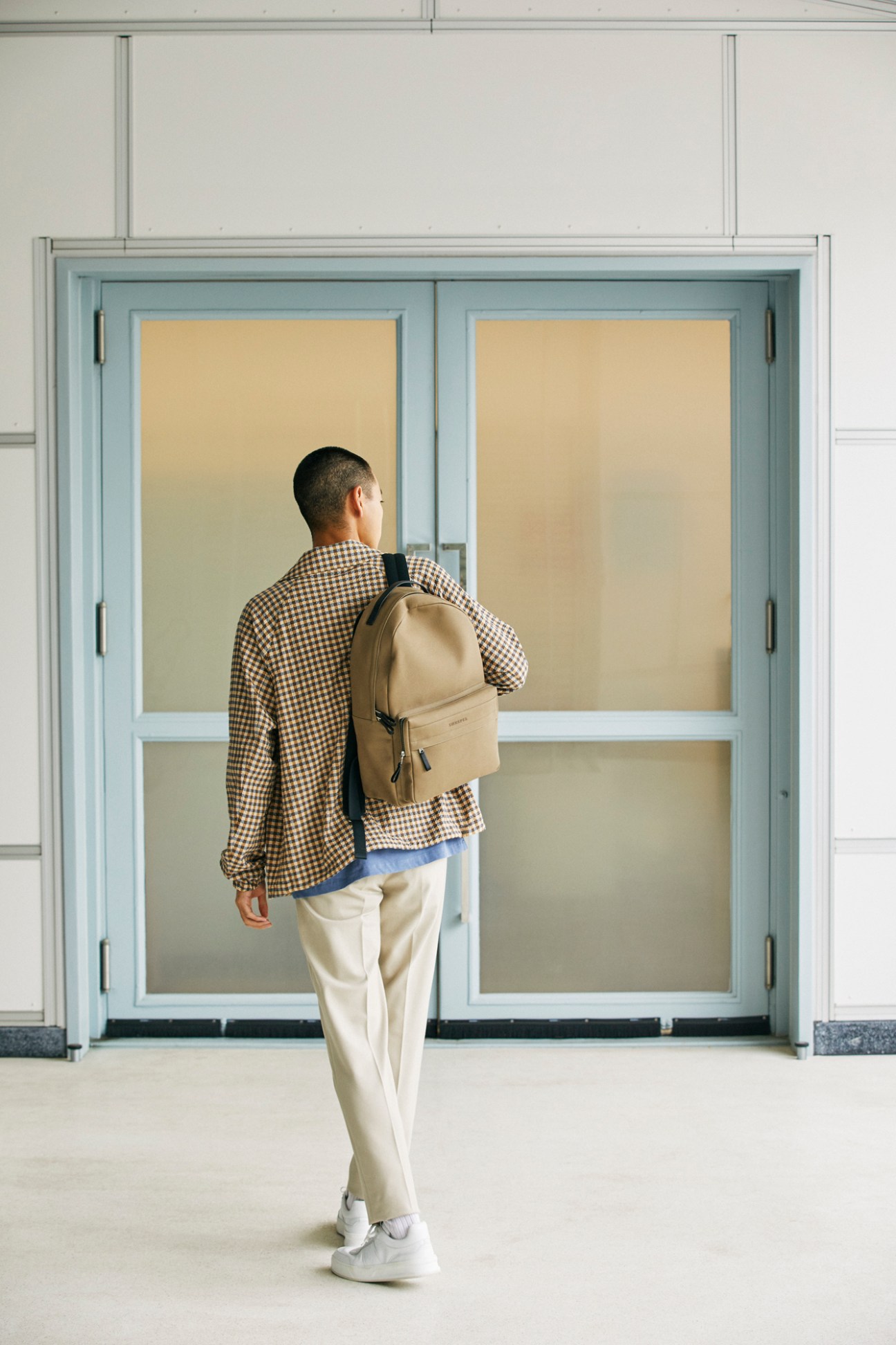 Man in checkered shirt and beige backpack standing before large glass doors in modern interior