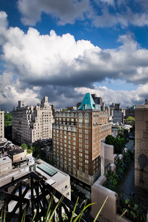Upper East Side Manhattan building with distinctive turquoise copper roof amid surrounding skyscrapers under blue sky with white clouds.