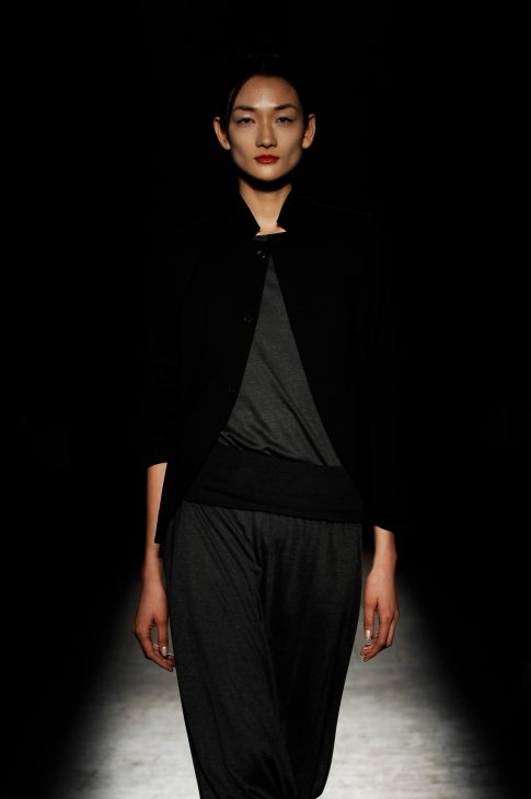 Model wearing black tailored dress with geometric panels against dark background with dramatic lighting