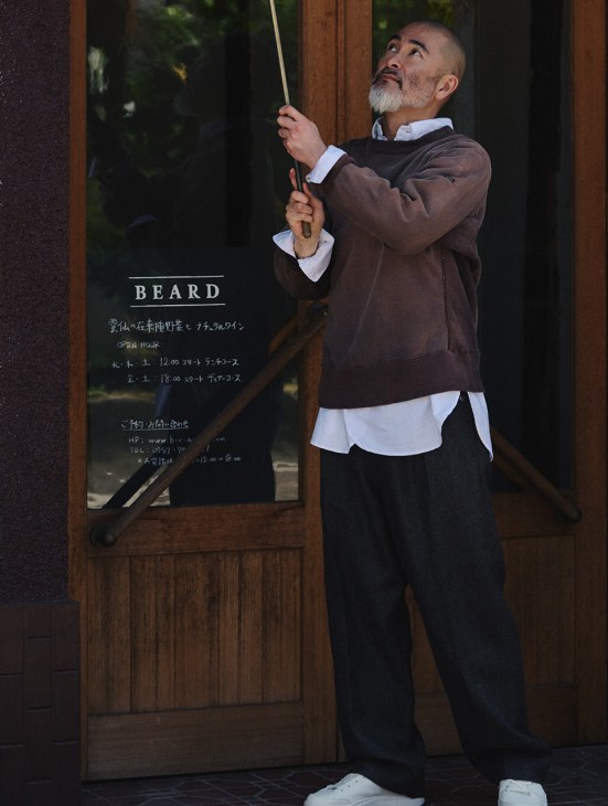 Man with grey beard wearing brown jumper, white shirt and black trousers standing outside a restaurant called Beard.