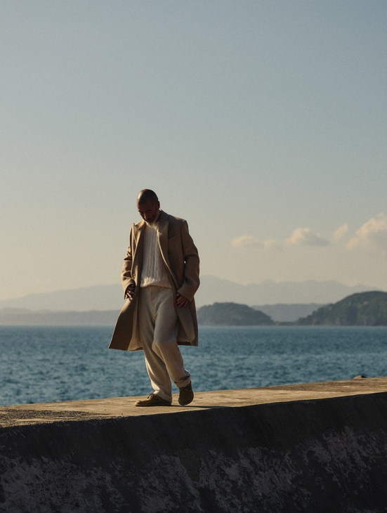 Chef Shinichiro Harakawa standing on a waterfront promenade wearing a camel coat, white jumper, and light trousers.