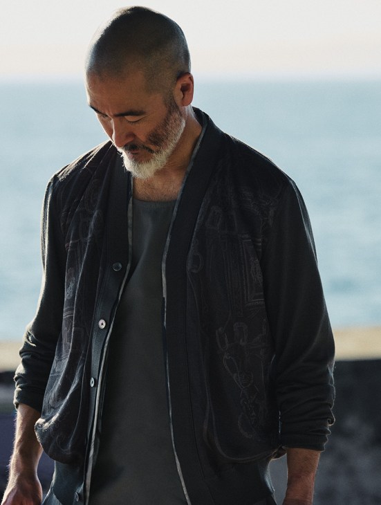 Chef Shinichiro Harakawa wearing a Hermès cardigan and vest, standing against a blurred seaside backdrop.
