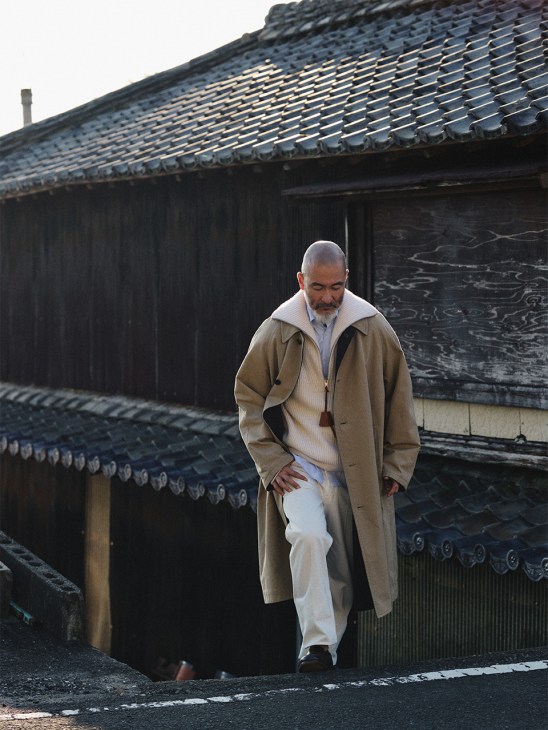 Chef Shinichiro Harakawa standing in front of traditional dark wooden Japanese architecture, wearing a beige Kaptain Sunshine coat over layered clothing.