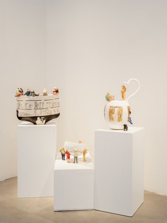 Three white ceramic vessels with gold accents displayed on gallery pedestals, featuring miniature figures.