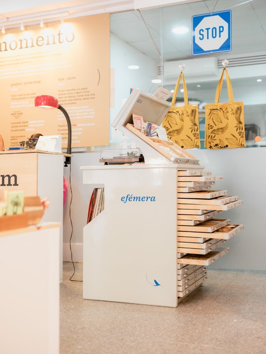 O Momento's ephemeral cabinet displaying yellow tote bags, paper stock, and a blue stop sign in a minimalist retail space.