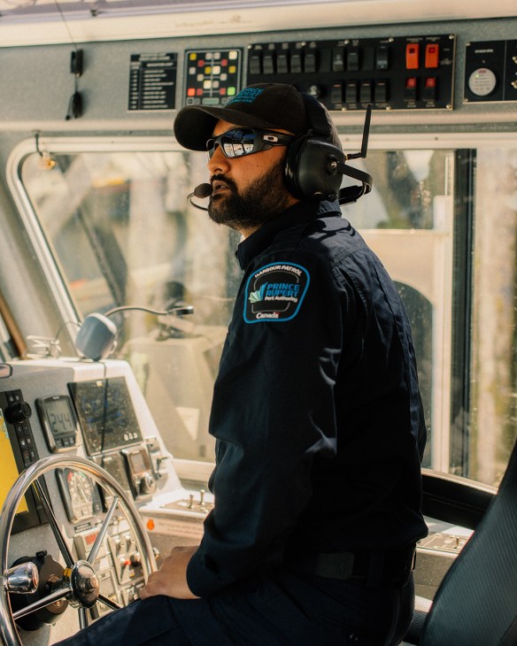 Harbour patrol vessel captain wearing headset and sunglasses in boat's wheelhouse with instrument panels behind.