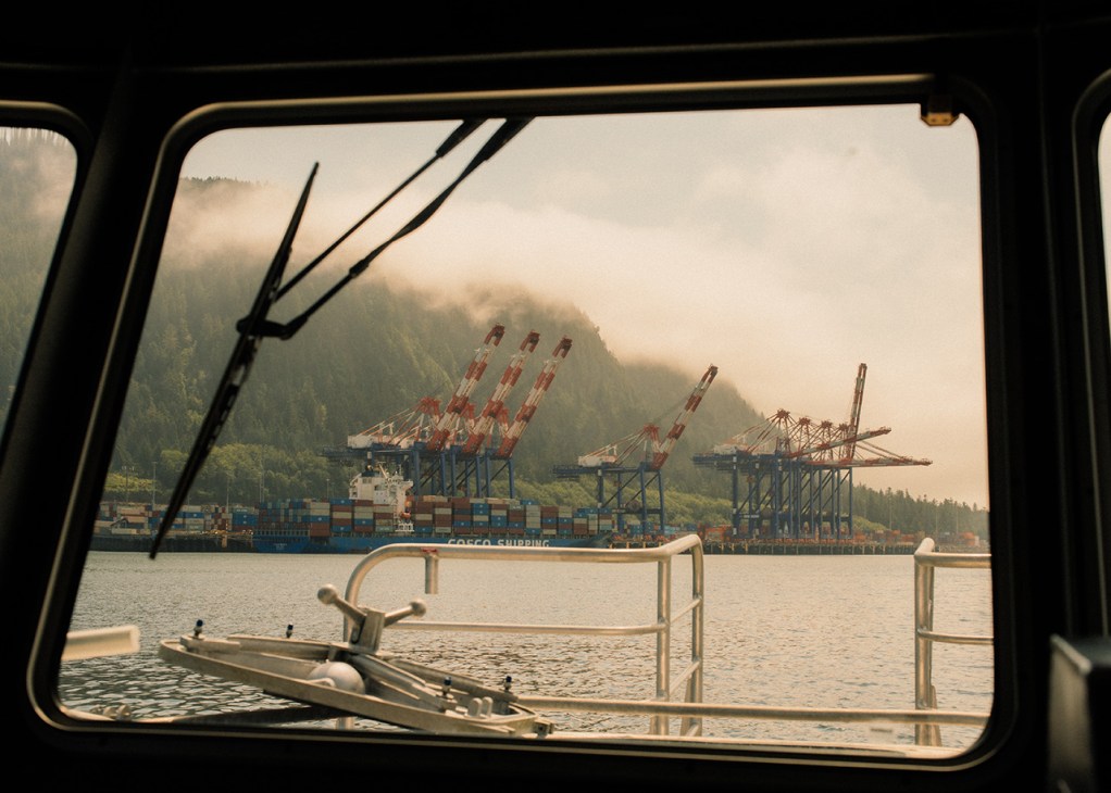 View from inside a vessel of a container ship docked at Fairview Container Terminal in Prince Rupert Harbour, with red cargo cranes and forested mountains in background.