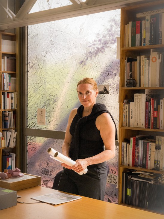 Tatiana Bilbao stands in her office holding architectural plans, surrounded by bookshelves with a colorful map visible behind her.