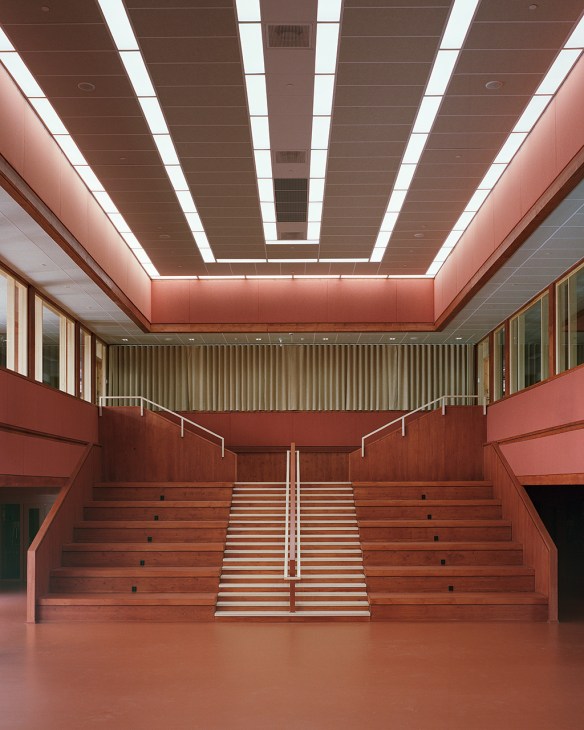 Modern school atrium with red-pink ceiling, wooden stairs, and natural lighting from linear ceiling panels.