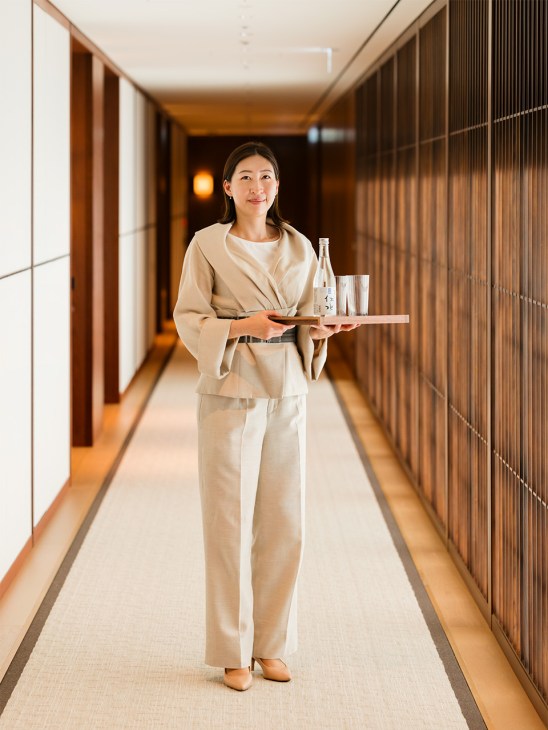 Woman in beige linen suit holding wooden tray with water bottle and glass in modern corridor.