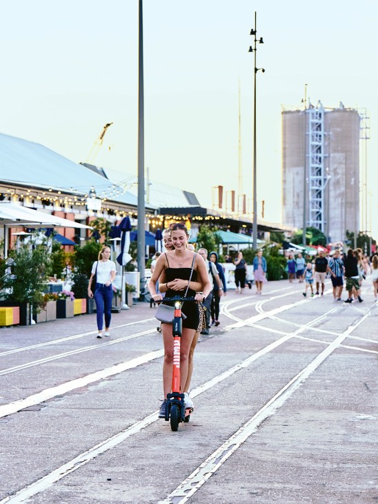 Woman riding a scooter along a pedestrian street at Auckland Waterfront with market stalls and restored industrial silos in the background.