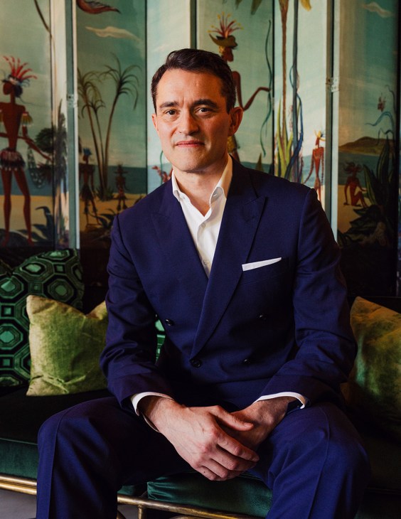 Francis Belin, CEO of Mytheresa, sits posed in a navy blazer against a jewel-toned backdrop with palm motifs.