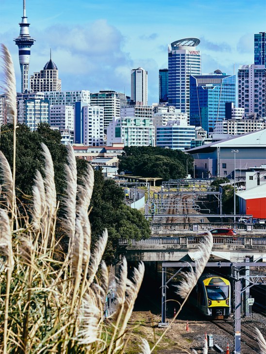 Auckland skyline with Sky Tower and modern buildings viewed from waterfront with train visible below.
