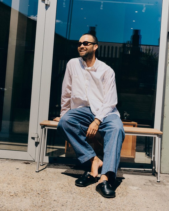Man wearing Paloceras sunglasses, white shirt and blue jeans sitting on wooden bench outside storefront.