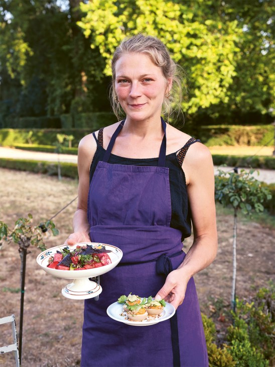 Sommelier Ioulia Gourieva holds two plated dishes in a garden setting, wearing a purple apron.