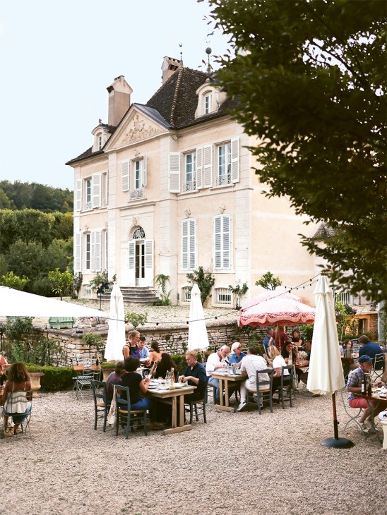 Diners seated at tables under white umbrellas in the garden courtyard of Ràde's pop-up restaurant, with a cream-colored château visible in the background.