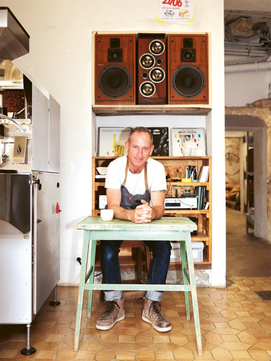 Matt McClune sits at a mint-green table in his Saint Romain Coffee roastery, with vintage wooden speakers mounted on the wall behind him.