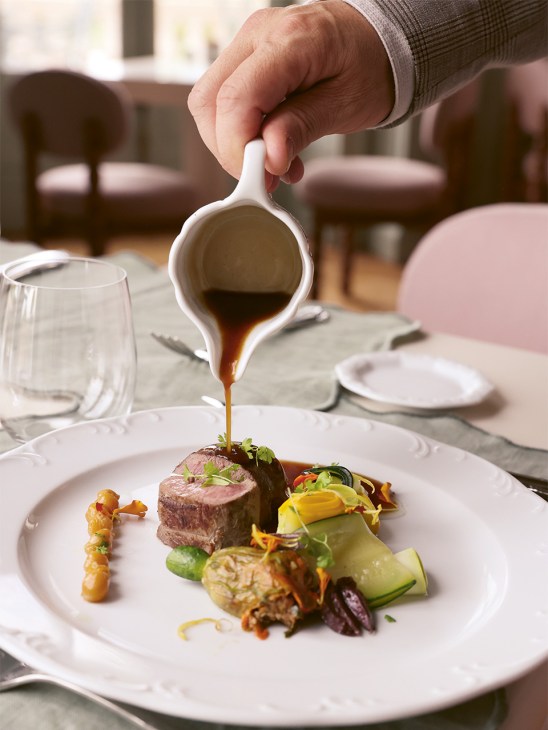 Hand pouring sauce over plated duck and vegetables at Château de Cîteaux restaurant.