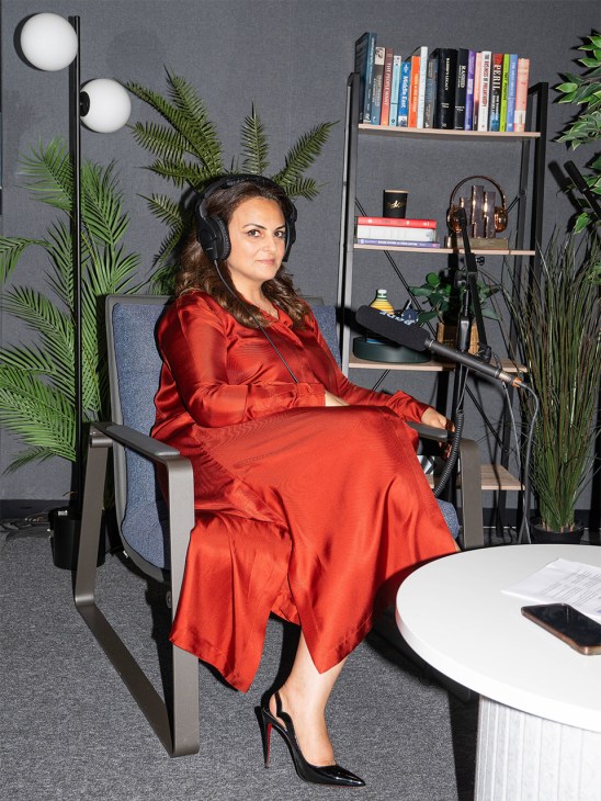 Woman in red dress wearing headphones sits in modern office chair with bookshelf and plants behind her