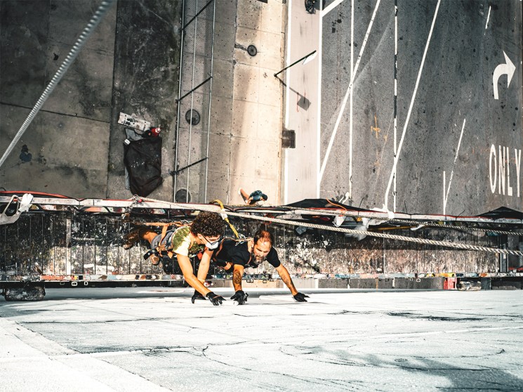 Two wall dogs suspended by safety lines paint a mural on the side of a building in New York City.