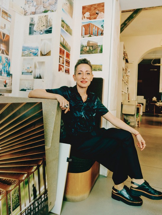 Architect Lina Ghotmeh poses in her Paris studio, surrounded by architectural photographs and mood boards on white walls.