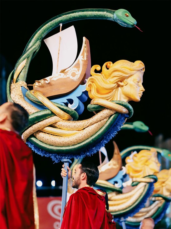 Ornate parade float featuring a green serpent, golden female head sculpture, white sail, and blue furry base.
