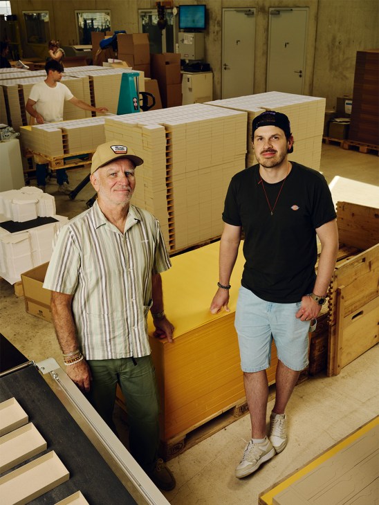 Bruno Sonderegger in striped shirt and cap stands with his son Dimitri in black shirt on the factory floor surrounded by stacked yellow cardboard boxes.