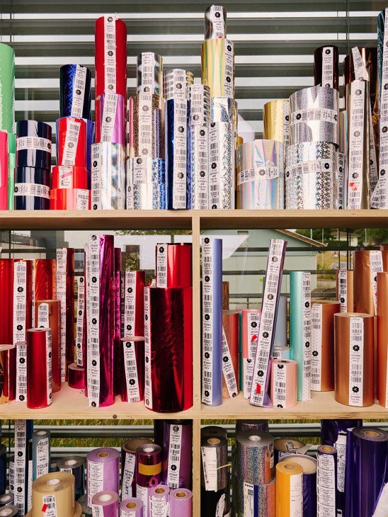 Shelving unit displaying colorful decorative foils and materials in rolls and tubes for packaging production.