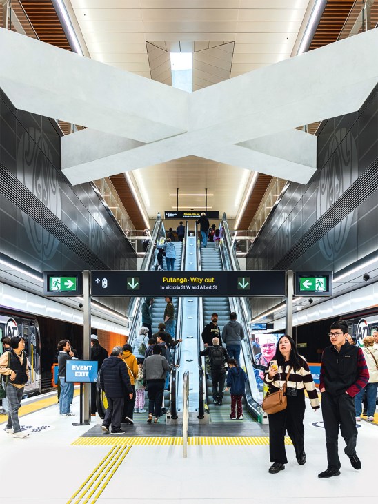 Te Waihorotiu underground metro station with symmetrical white ceiling beams, escalators, and passengers moving through the platform
