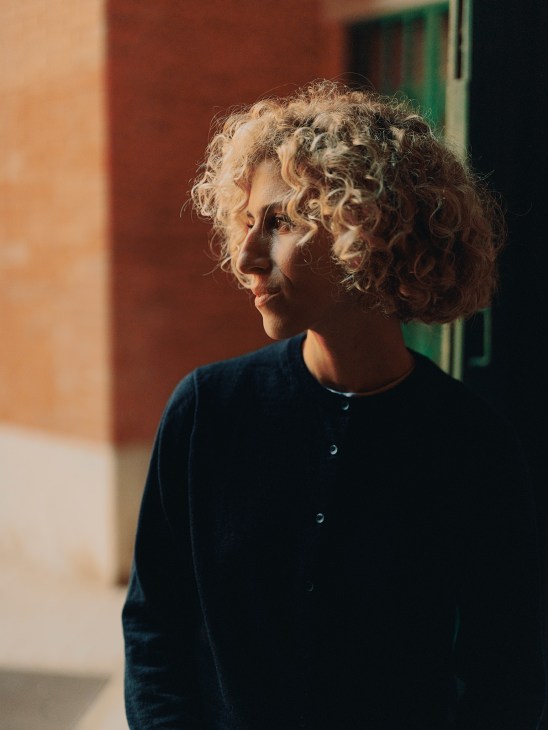 Woman with curly blonde hair wearing dark blue shirt looking to the side against colorful building backdrop