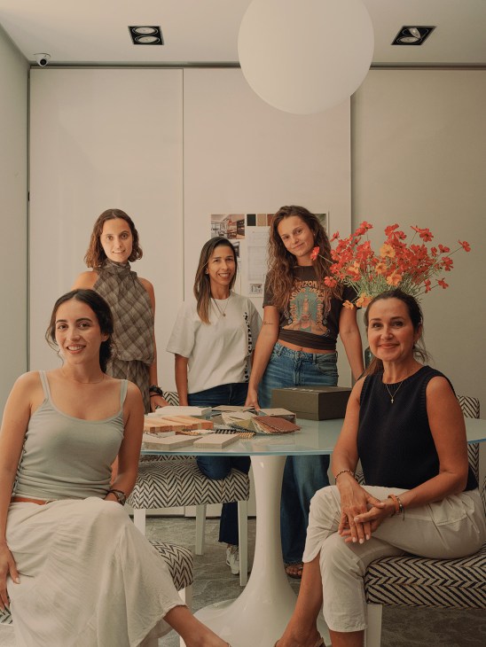 Five women pose in Laura Yerpes' interior design studio with material samples on a table and red flowers.