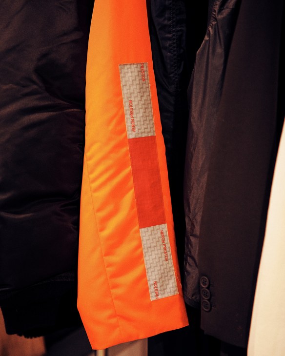 Close-up of an orange Heron Preston jacket sleeve featuring reflective striping and branded patches.