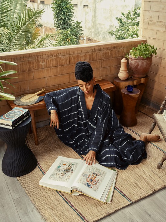 Jeanne Autran-Edorh sits on a wooden floor in her Lomé home, wearing a navy tie-dye patterned outfit, studying an open book with architectural drawings.
